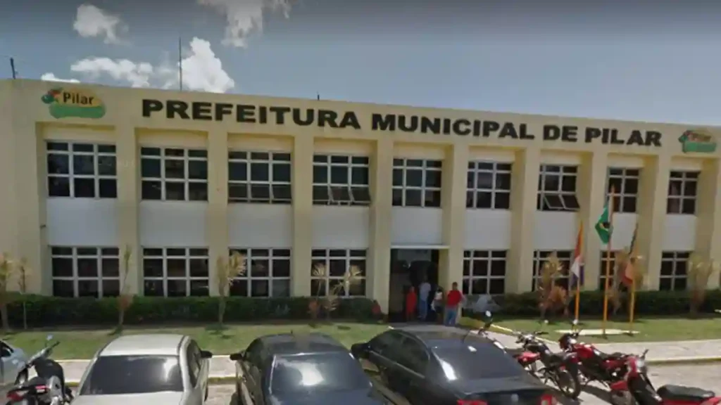 Fachada da Prefeitura Municipal de Pilar, com carros e motos estacionados em frente e mastros com bandeiras na entrada