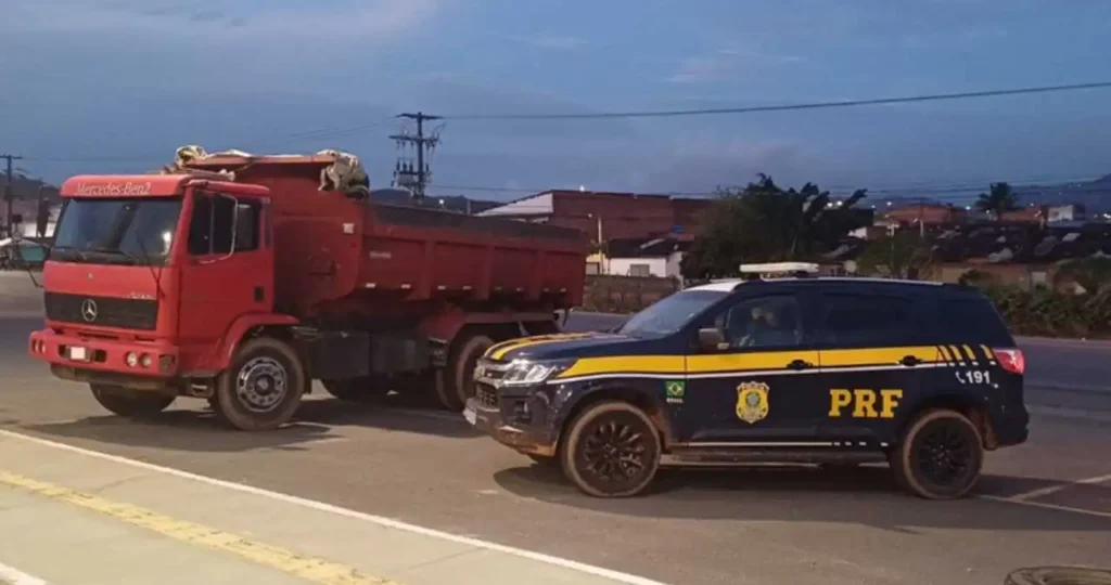 Em um acostamento de asfalto sob um céu de fim de tarde, uma viatura da Polícia Rodoviária Federal (PRF) — modelo SUV preto e azul com detalhes amarelos — está estacionada à frente de um caminhão caçamba vermelho da marca Mercedes-Benz. A viatura e as rodas do caminhão estão sujas de lama.