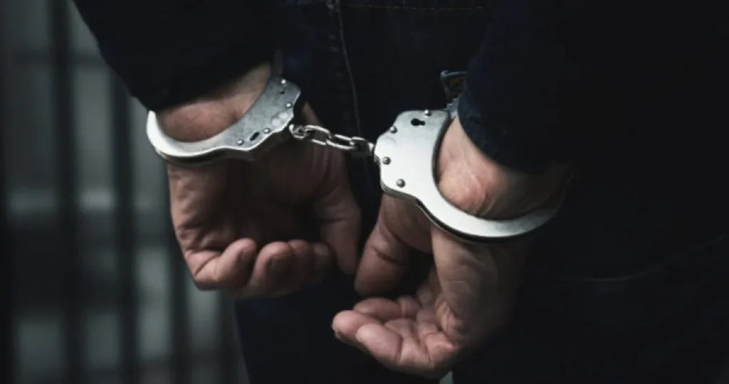 Close-up das costas de um homem vestindo calça jeans escura, com as mãos algemadas para trás. Ao fundo, de forma desfocada, percebe-se a estrutura de grades de uma cela ou unidade policial.