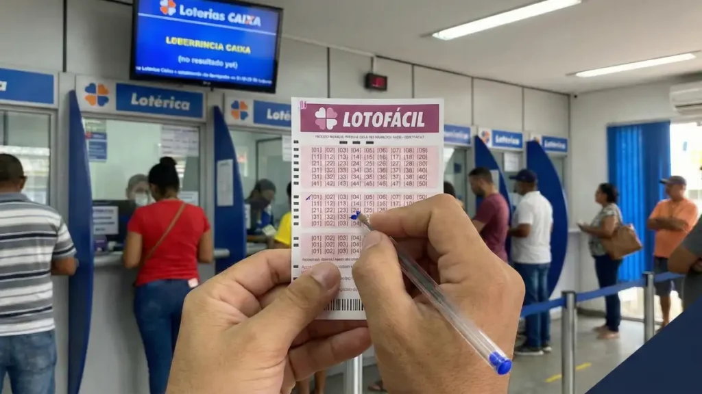 Apostador segura e marca com caneta um volante da Lotofácil dentro de uma lotérica; ao fundo há guichês de atendimento, fila de clientes e um monitor das Loterias CAIXA.