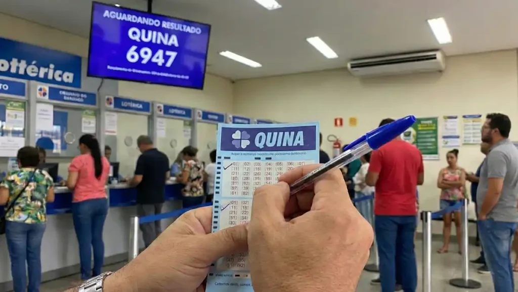 Apostador segura um volante da Quina e uma caneta dentro de uma lotérica; ao fundo, clientes aguardam atendimento e um painel informa “Aguardando resultado – Quina 6947”.