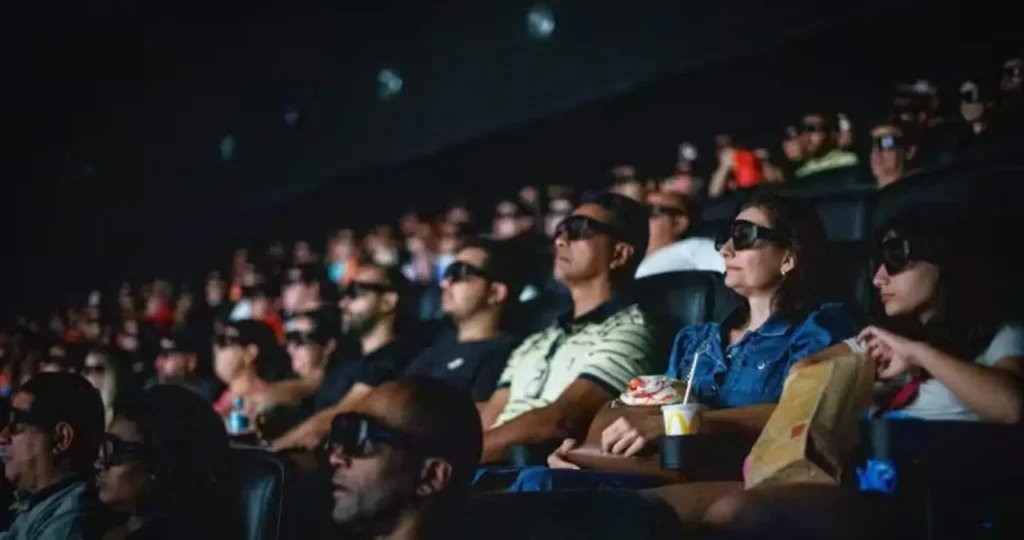 Fileiras de espectadores sentados em poltronas pretas de uma sala de cinema escura. Eles vestem óculos 3D pretos e olham fixamente para a frente. Uma mulher em destaque segura um saco de pipoca de papel e há um copo de refrigerante no suporte da poltrona.