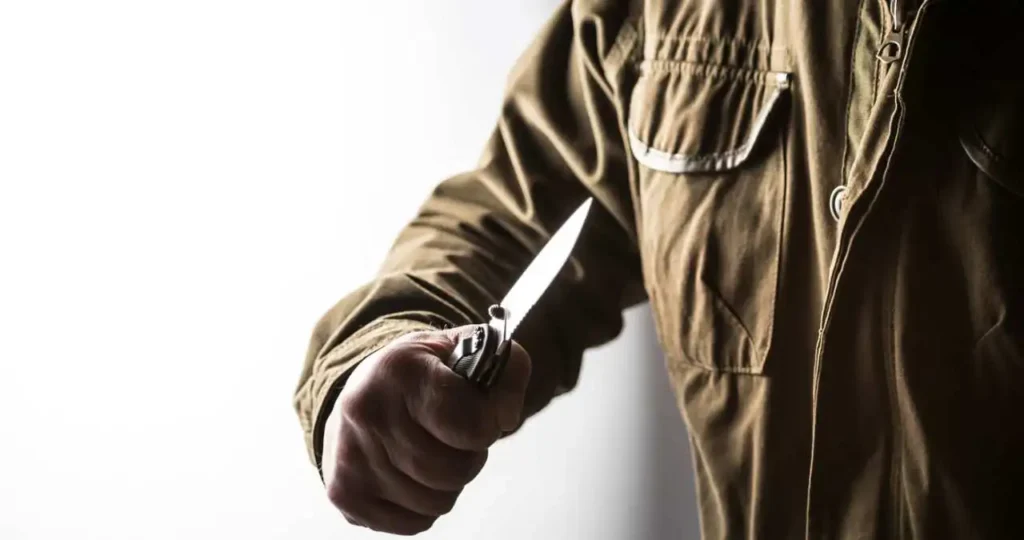 Close-up lateral de uma pessoa vestindo uma jaqueta de lona marrom, segurando um canivete preto aberto com a lâmina prateada apontada para a frente. O fundo é branco e bem iluminado, criando um forte contraste com a silhueta da pessoa e da arma branca.