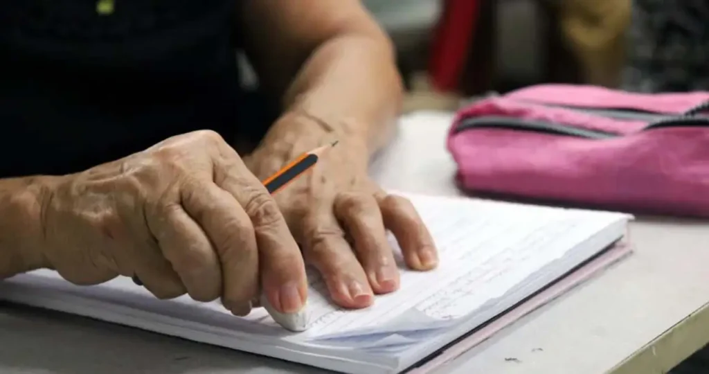 Close-up das mãos enrugadas de uma pessoa idosa sobre um caderno pautado em uma mesa de escola. Com a mão direita, a pessoa usa uma borracha branca para apagar algo escrito a lápis. Ao lado, há um estojo rosa e a mão esquerda apoia o caderno.