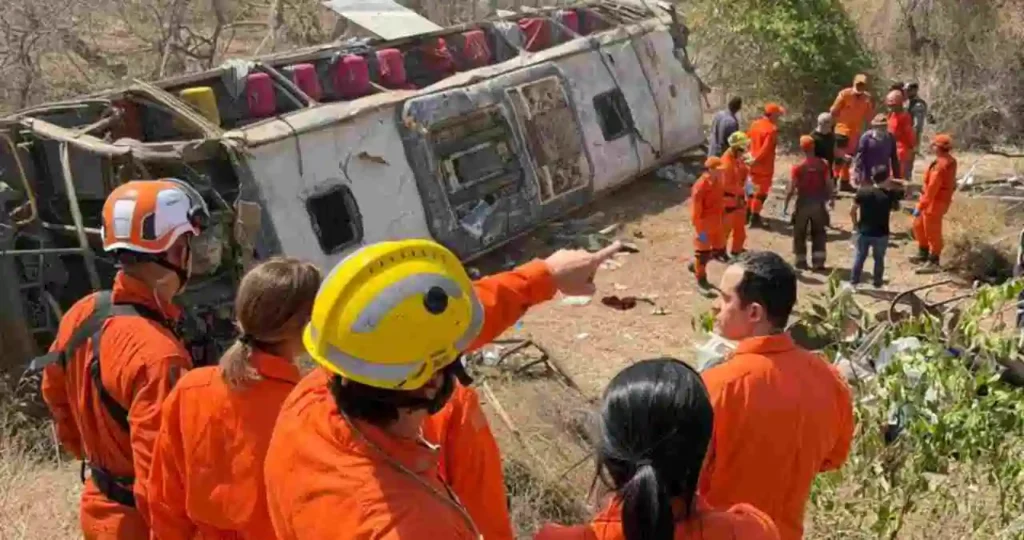 No alto de uma ribanceira, quatro socorristas do Corpo de Bombeiros em uniformes laranja — três homens e uma mulher — observam um ônibus de turismo branco capotado lá embaixo. O ônibus está tombado lateralmente com as rodas para cima e o teto destruído. Outras equipes de resgate circulam ao redor do veículo no terreno seco.