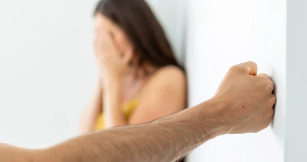 Em um ambiente interno de paredes brancas, o braço peludo de um homem está em primeiro plano, com o punho fechado contra a parede. Ao fundo, uma mulher com cabelos escuros está sentada, desfocada, cobrindo o rosto com as mãos em um gesto de angústia.